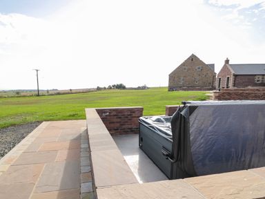 An outdoor hot tub with a partially open cover on a stone patio near brick buildings at Byre Cottage in Embleton