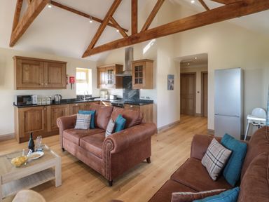 An open plan kitchen and living room with wooden beams on the ceiling brown sofas and wooden cabinets at Smithy Cottage in Embleton