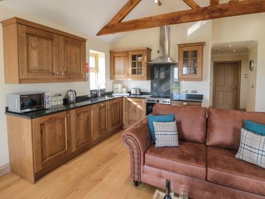 A kitchen with wooden cabinets a microwave kettle toaster stove and a brown sofa with cushions at Smithy Cottage in Embleton