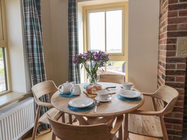 A dining area with a round wooden table set with white dishes and a glass vase of purple flowers at Smithy Cottage Embleton
