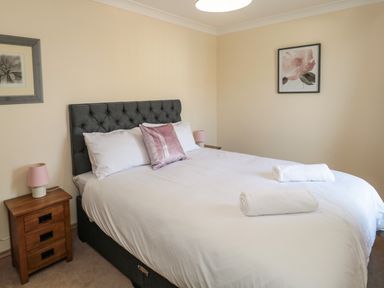 A bedroom with a double bed white bedding and two rolled towels a wooden bedside table with a lamp and framed pictures on the walls at Smithy Cottage in Embleton
