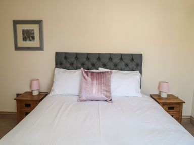 A bedroom with a bed with a grey headboard pink pillow and two wooden bedside tables with pink lamps at Smithy Cottage in Embleton