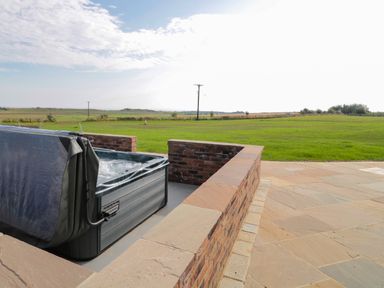 An outdoor hot tub with a partially removed cover on a stone patio overlooking a grassy field at Smithy Cottage in Embleton