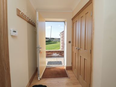 An entryway with an open door showing a green lawn outside a wooden closet and coat hooks on the wall at Bothy Cottage in Embleton