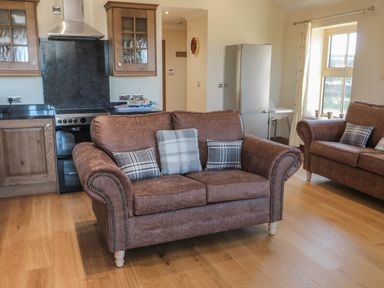 A living room with two brown sofas and a kitchen with wooden cabinets and a stainless steel fridge at Bothy Cottage in Embleton