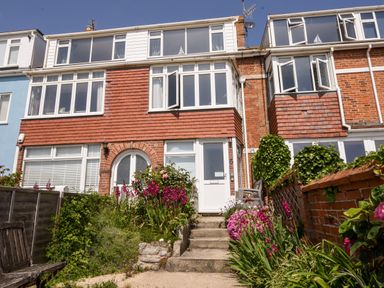 A front view of a three-story brick house with stairs leading to a white door and flowers on both sides at The Penthouse @ No 5 in Lyme Regis