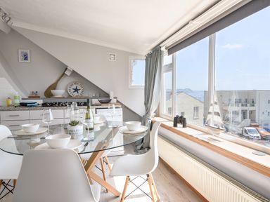 A kitchen and dining area with a glass table set for four near large windows overlooking buildings and the sea at The Penthouse @ No 5 in Lyme Regis