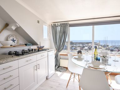 A kitchen and dining area with white cabinets a gas stove a glass dining table set with plates bowls and glasses and a window overlooking a harbor at The Penthouse @ No 5 in Lyme Regis