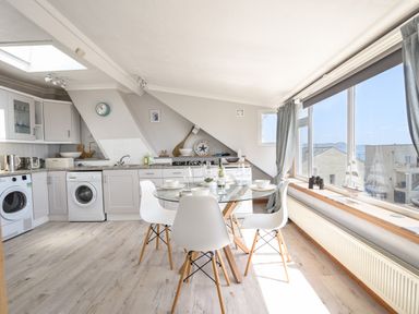 A kitchen with white cabinets a round glass dining table and white chairs by a large window at The Penthouse @ No 5 in Lyme Regis