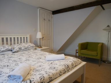 A bedroom with a bed with floral bedding two rolled towels a white bedside table with lamp a green armchair and a floor lamp at Pebble Reef in Easton Portland