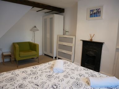 A bedroom with a patterned bedspread two rolled towels green armchair white wardrobe chest of drawers black fireplace and wall lamp at Pebble Reef Easton Portland