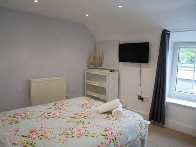 A bedroom with a bed covered in floral bedding a chest of drawers a mirror a TV on the wall and a window with curtains at Pebble Reef in Easton Portland