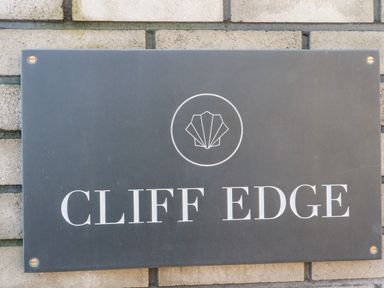 A black rectangular sign with a shell logo and the text Cliff Edge mounted on a brick wall at 33 Cliff Edge in Newquay
