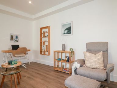A room with a desk and chair a beige armchair with ottoman and wooden shelves at 33 Cliff Edge in Newquay