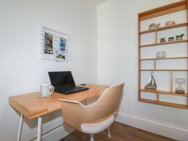 A home office with a laptop on a wooden desk and a wooden chair next to a wall-mounted wooden shelf at 33 Cliff Edge in Newquay