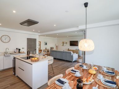 An open-plan kitchen and dining area with a wooden dining table set with plates and cups a kitchen island with a basket of fruit and a living area with a grey sofa at 33 Cliff Edge in Newquay