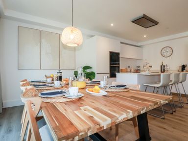 A dining table set with plates cups and a jug of orange juice next to a kitchen island with bar stools and white cabinets at 33 Cliff Edge in Newquay