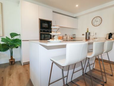 A kitchen with white cabinets a kitchen island with stools a plant and a wall clock at 33 Cliff Edge in Newquay