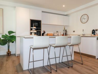 A kitchen with white cabinets a kitchen island with three white bar stools a wall clock and a potted plant at 33 Cliff Edge in Newquay