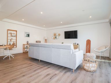 A living room with a gray sofa facing a wall-mounted TV a wicker basket on wooden floor and a small chair at 33 Cliff Edge in Newquay