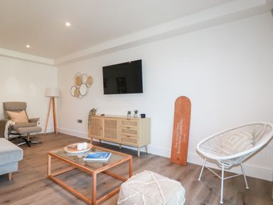 A living room with a wall mounted TV a wooden sideboard a gray armchair a white woven chair a coffee table and a floor lamp at 33 Cliff Edge in Newquay