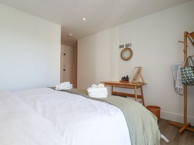 A bedroom with a bed with white linens green blanket and folded towels a wooden desk with a mirror and bottles a coat rack with a towel and bag and a wicker basket on the floor at 33 Cliff Edge in Newquay