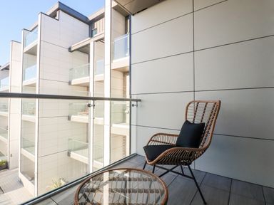 A balcony with a wicker chair and glass table overlooking a modern building at 33 Cliff Edge in Newquay