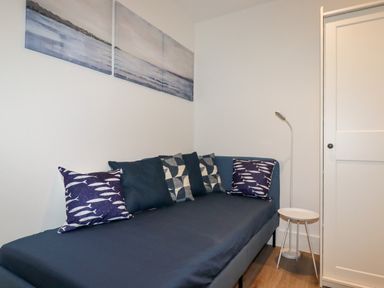 A small bedroom with a navy blue bed and several pillows including fish patterned ones next to a white wardrobe and a small white side table with a floor lamp at 33 Cliff Edge in Newquay