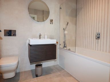 A bathroom with a toilet a sink with a cabinet a round mirror a trash bin and a bathtub with a shower at 33 Cliff Edge in Newquay
