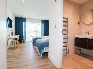 A bedroom with two beds covered in blue throws a desk and chair a wall-mounted tv and a bathroom with a round mirror and sink at 33 Cliff Edge in Newquay