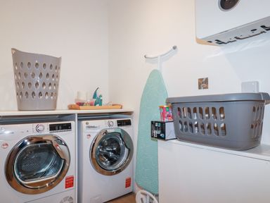 A laundry room with a washing machine and dryer two laundry baskets an ironing board and various laundry supplies at 33 Cliff Edge in Newquay