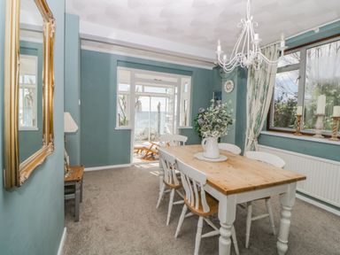 A dining room with a wooden table and white chairs a large mirror on the wall and windows overlooking the outside at Raven Cottage in Downderry