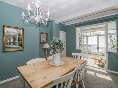 A dining room with a wooden table and white chairs overlooking water through glass sliding doors at Raven Cottage in Downderry