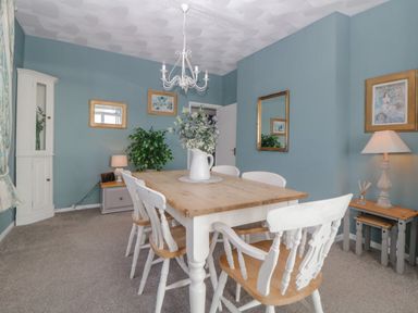 A dining room with a wooden table and white chairs a chandelier and framed pictures on blue walls at Raven Cottage in Downderry
