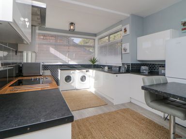 A kitchen with white cabinets black countertops a washing machine a microwave and a dining area at Raven Cottage in Downderry