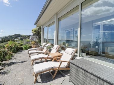 A patio with cushioned lounge chairs and a small table outside large glass windows at Raven Cottage in Downderry