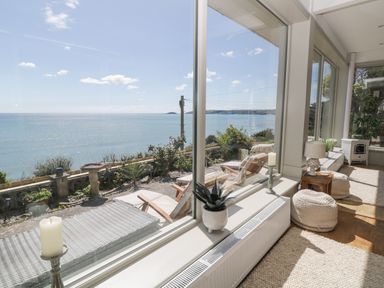 A sunroom with large windows overlooking the sea with chairs plants candles and poufs at Raven Cottage in Downderry