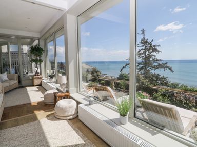 A sunroom with large windows overlooking the ocean with chairs a sofa plants and soft ottomans at Raven Cottage in Downderry