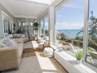 A sunroom with beige sofas, pillows, plants, poufs, and large windows overlooking the sea at Raven Cottage in Downderry