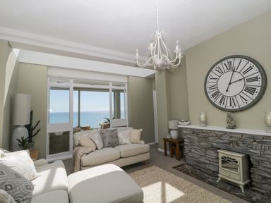 A living room with sofas a large clock on the wall and a sea view through glass doors at Raven Cottage in Downderry