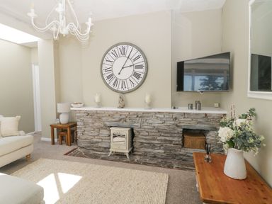 A living room with a stone fireplace a large clock wall mounted tv and wooden tables at Raven Cottage in Downderry