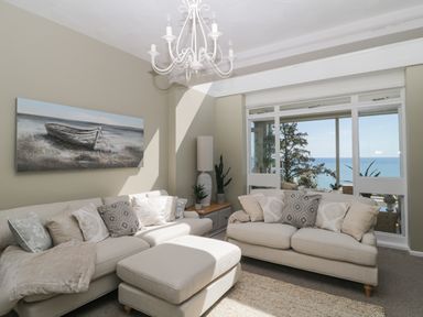 A living room with two beige sofas cushions a large window with sea view and a painting of a boat on the wall at Raven Cottage in Downderry