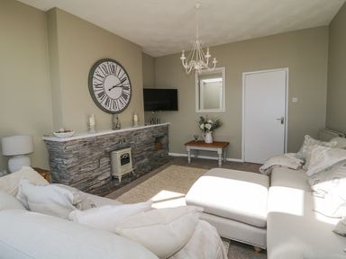 A living room with a large clock above a stone fireplace and a beige sofa at Raven Cottage in Downderry