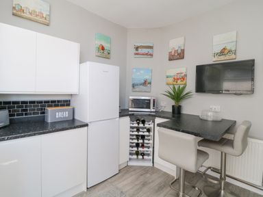 A kitchen corner with white cabinets a refrigerator a microwave and a wine rack with bar stools and a wall mounted television at Raven Cottage in Downderry