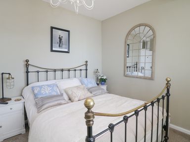 A bedroom with a metal framed bed white bedding blue and beige pillows two bedside tables with lamps a wall mirror and a framed picture at Raven Cottage in Downderry