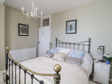 A bedroom with a metal bed frame white bedding pillows a white nightstand with a lamp and flowers paintings on the walls Raven Cottage Downderry