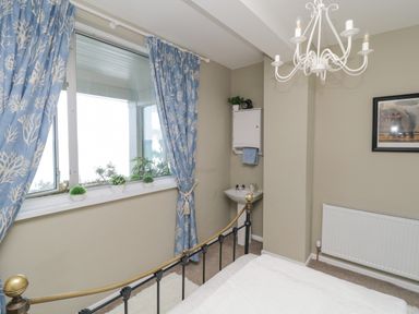 A bedroom with blue curtains on a large window a metal bed frame a white sink and cupboard and a radiator on the wall at Raven Cottage in Downderry