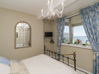 A bedroom with a bed a wall mirror a small wall-mounted TV and window with blue patterned curtains overlooking the sea at Raven Cottage in Downderry