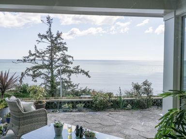 A patio with wicker chair and table overlooking the ocean with plants around at Raven Cottage in Downderry