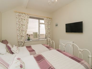 A bedroom with two single beds with floral and checkered bedding a window with floral curtains a wall-mounted television and a small framed picture at Raven Cottage in Downderry
